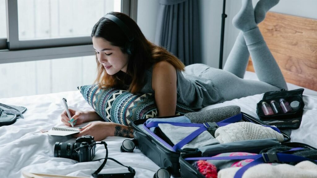 Woman lying on bed with headphones, writing in notebook, and packing suitcase.