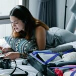 Woman lying on bed with headphones, writing in notebook, and packing suitcase.