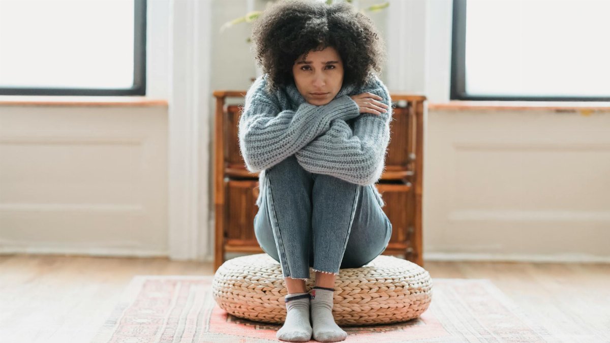 Full body of sad African American female looking at camera while sitting on rattan mat in light room at home