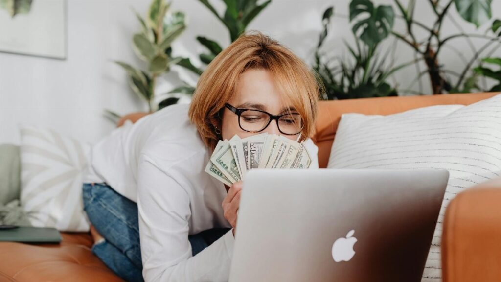 Woman holding cash while using a laptop on a cozy sofa at home, conveying financial success.