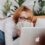 Woman holding cash while using a laptop on a cozy sofa at home, conveying financial success.