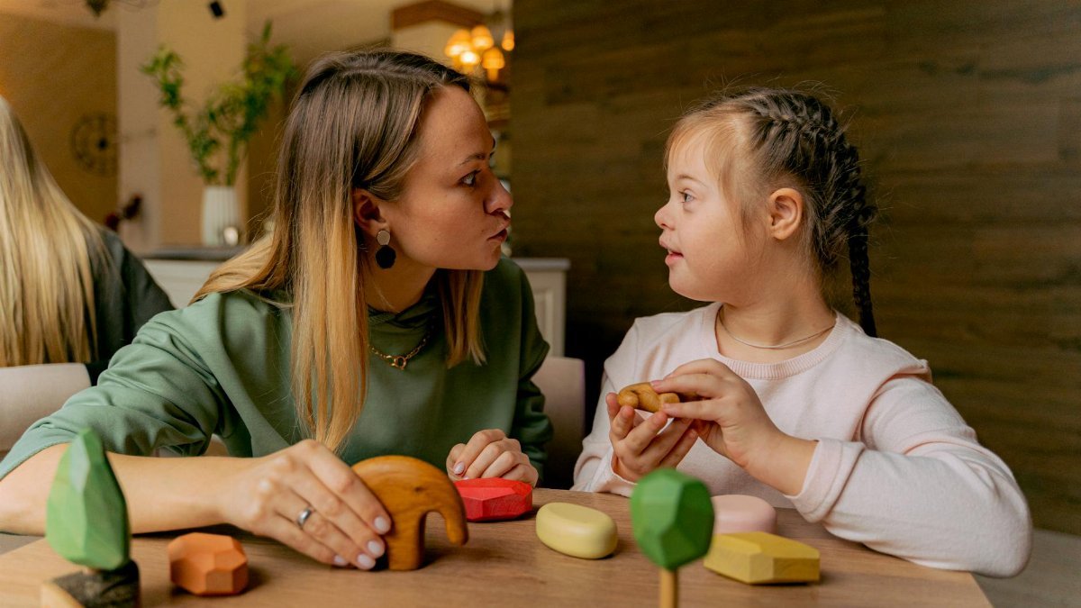 An adult woman and a child with Down syndrome engaging in playful interaction with wooden toys indoors.