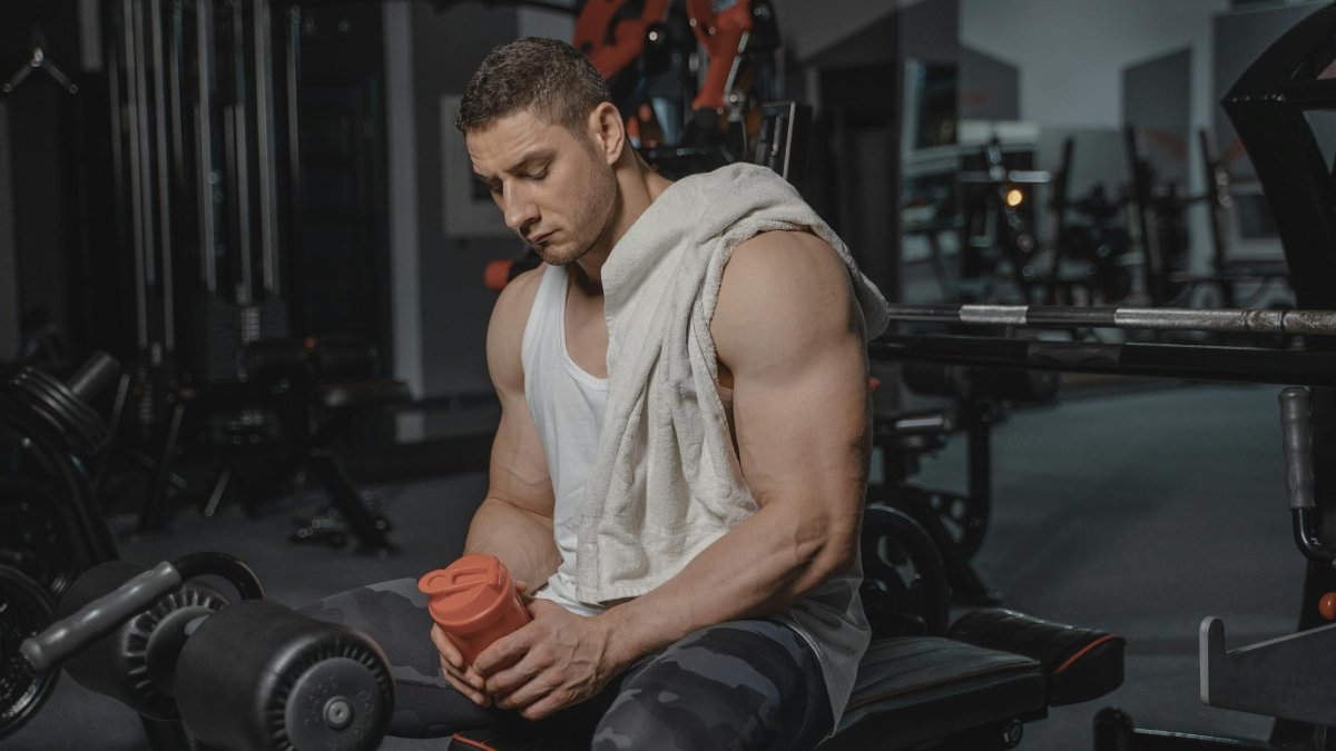 Muscular man sits in gym holding protein shaker after a workout, towel on shoulder.
