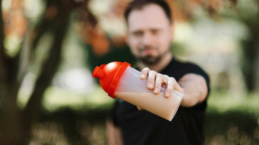 Man presenting a protein shake in a plastic tumbler against a blurred nature background.