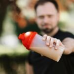 Man presenting a protein shake in a plastic tumbler against a blurred nature background.