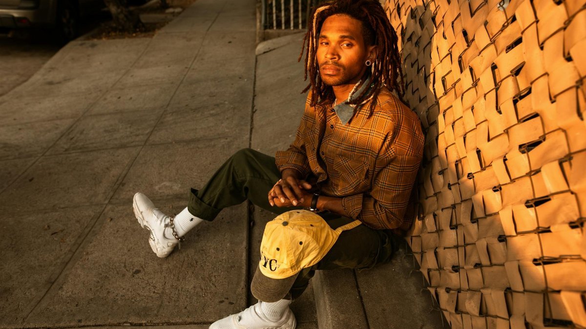 A man with dreadlocks sits on the sidewalk, bathed in golden sunset light.