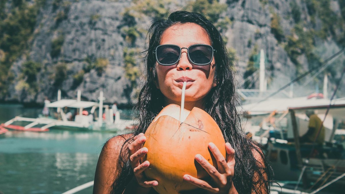 A woman in sunglasses enjoying a fresh coconut drink by the water, with boats and mountains in the background.