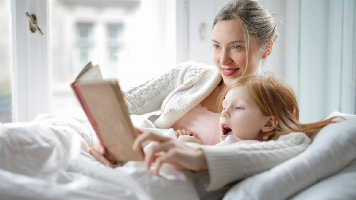 Cheerful young woman hugging cute little girl and reading book together while lying in soft bed in light bedroom at home in daytime
