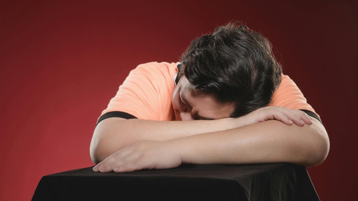 An adult man rests his head on a table, symbolizing fatigue or contemplation, set against a rich red background.