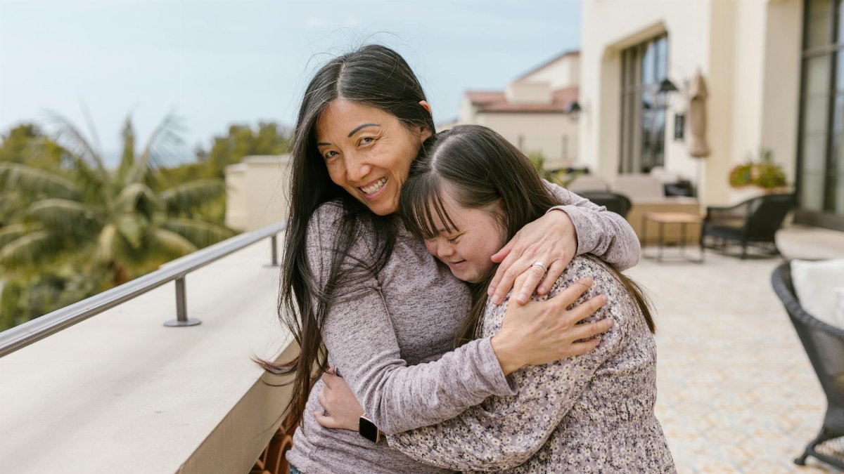 Mother and daughter with Down syndrome share a warm embrace outdoors on a sunny day.