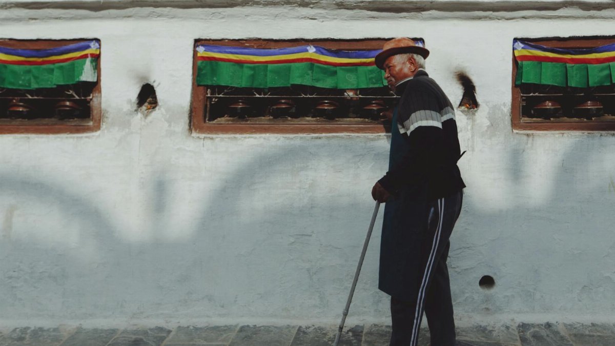 Senior man walking with a cane past a vibrant wall adorned with prayer wheels.