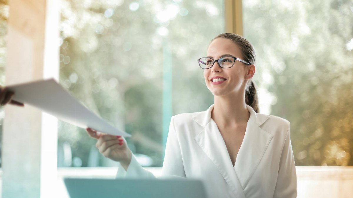 Low angle of successful female executive manager in classy style sitting at table with laptop in contemporary workplace and passing documents to colleague