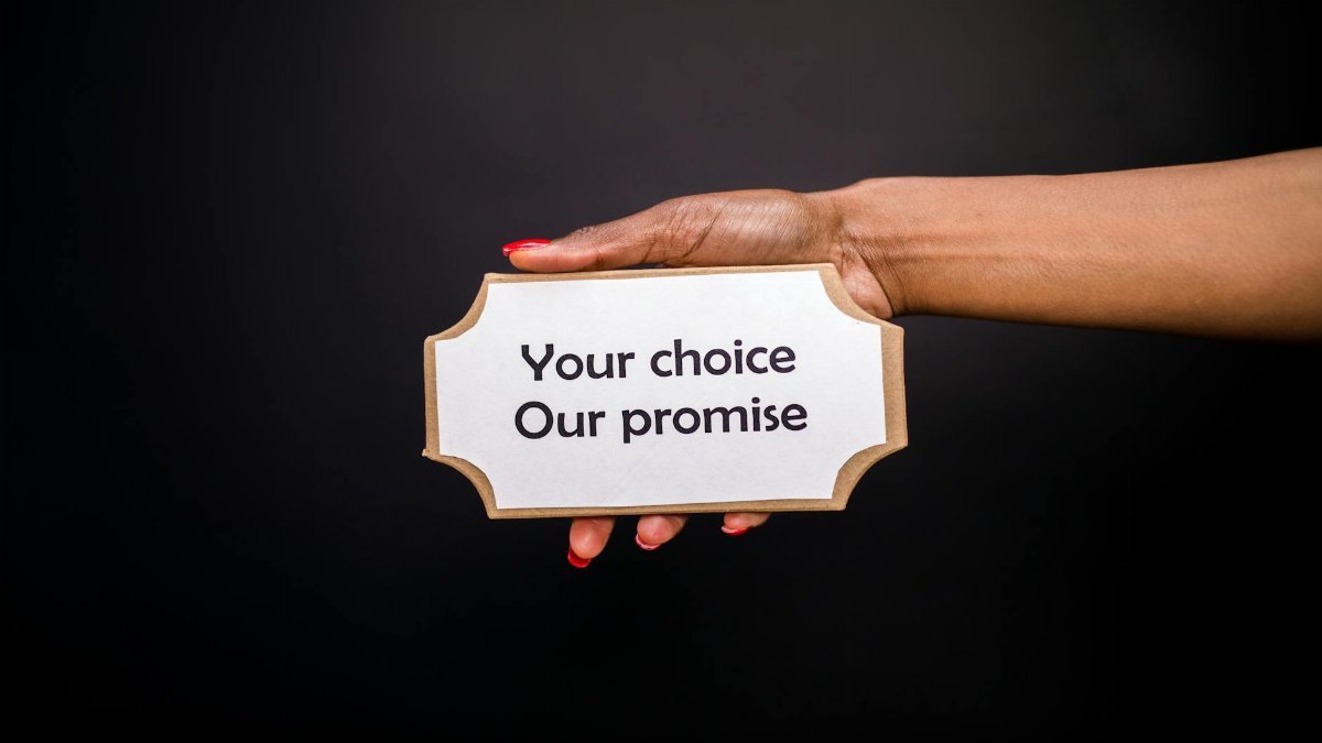 A hand holds a sign saying 'Your choice, Our promise' against a dark background.