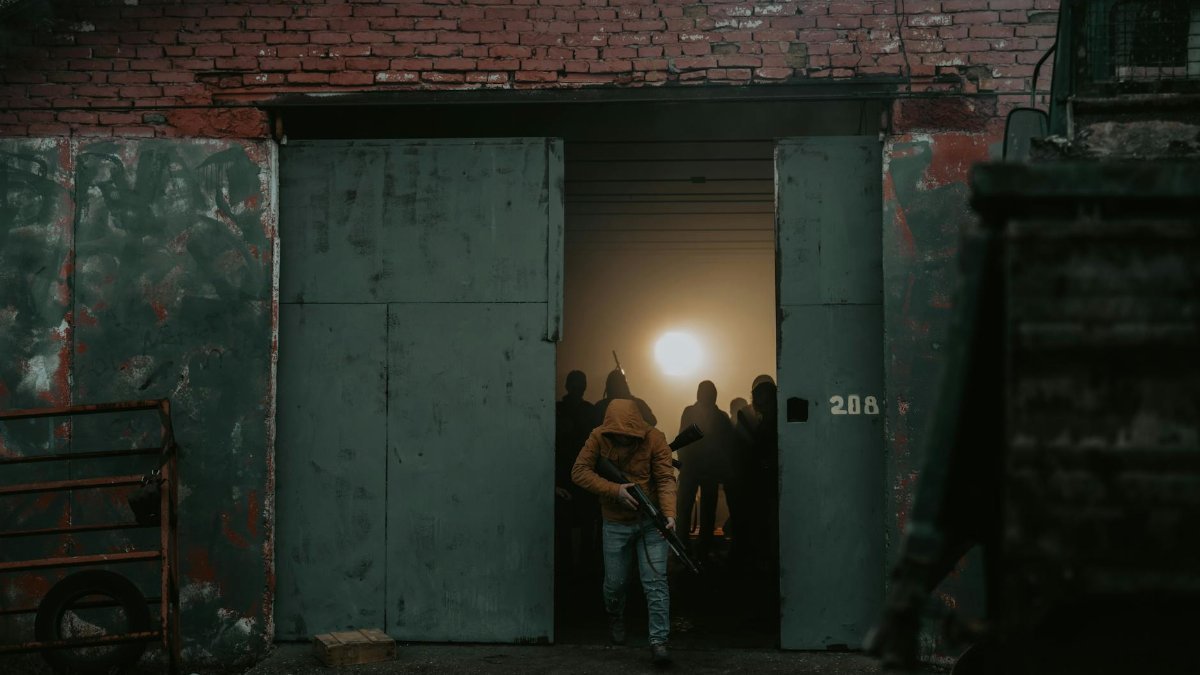 A person in a hoodie with a firearm exits a dark building at dusk, creating a dramatic and tense atmosphere.