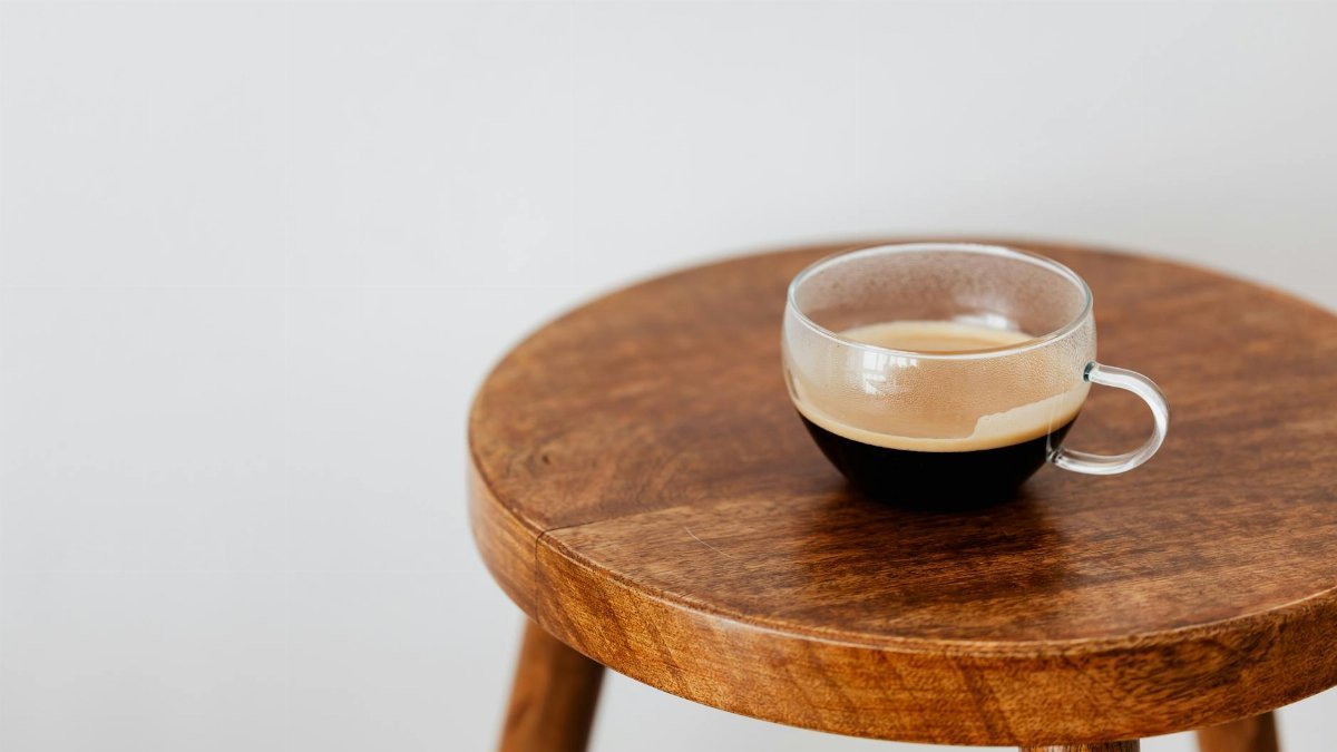 Wide cup of fresh black coffee placed on small brown wooden table near white wall