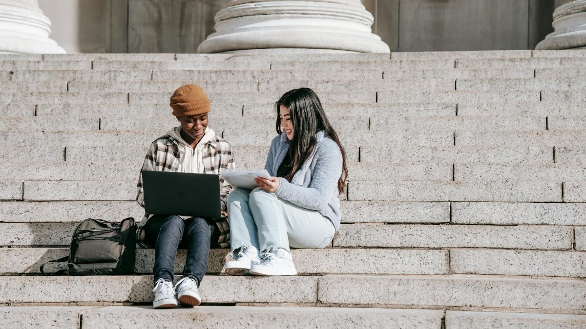 Full length cheerful young multiracial female students sitting on stairs with laptop and tablet while doing home task together