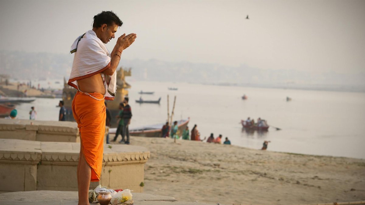 Devout man in orange robe praying on the riverbank at sunrise, a serene and spiritual moment.