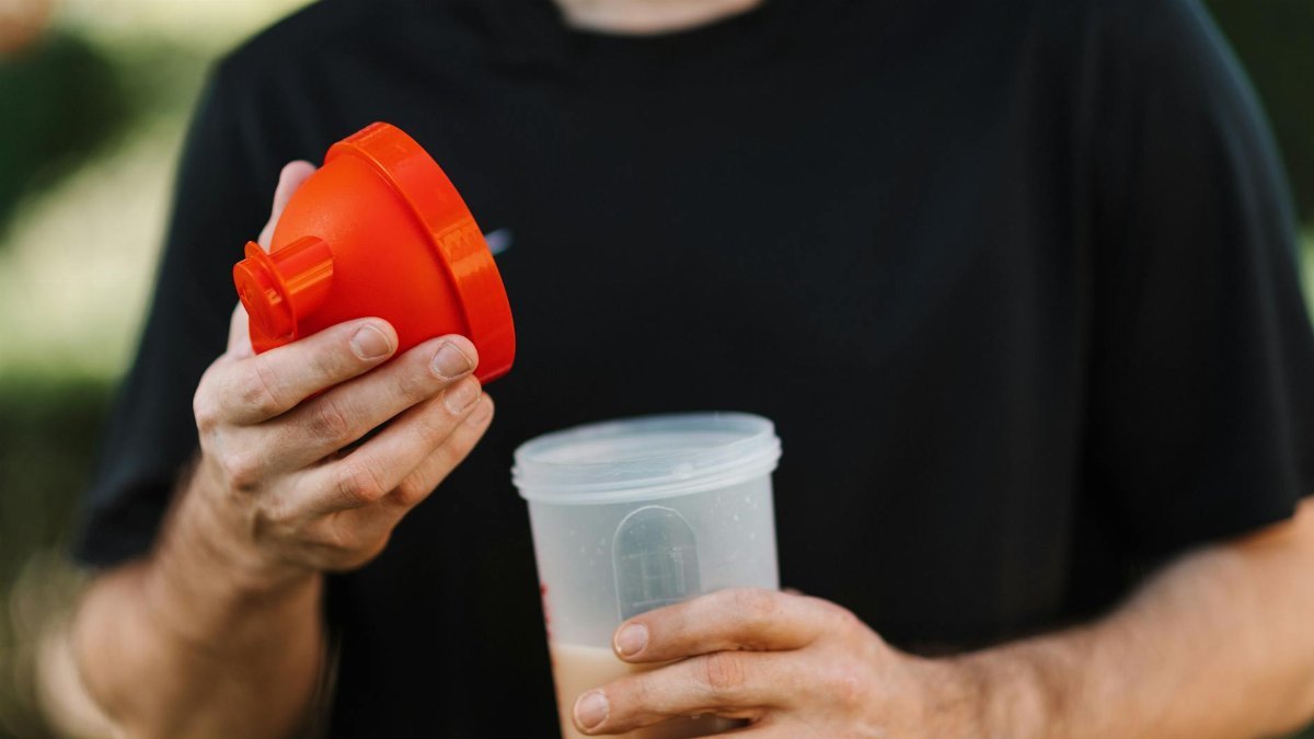 A man in a black shirt holds a protein shake tumbler with an orange lid, outdoors.