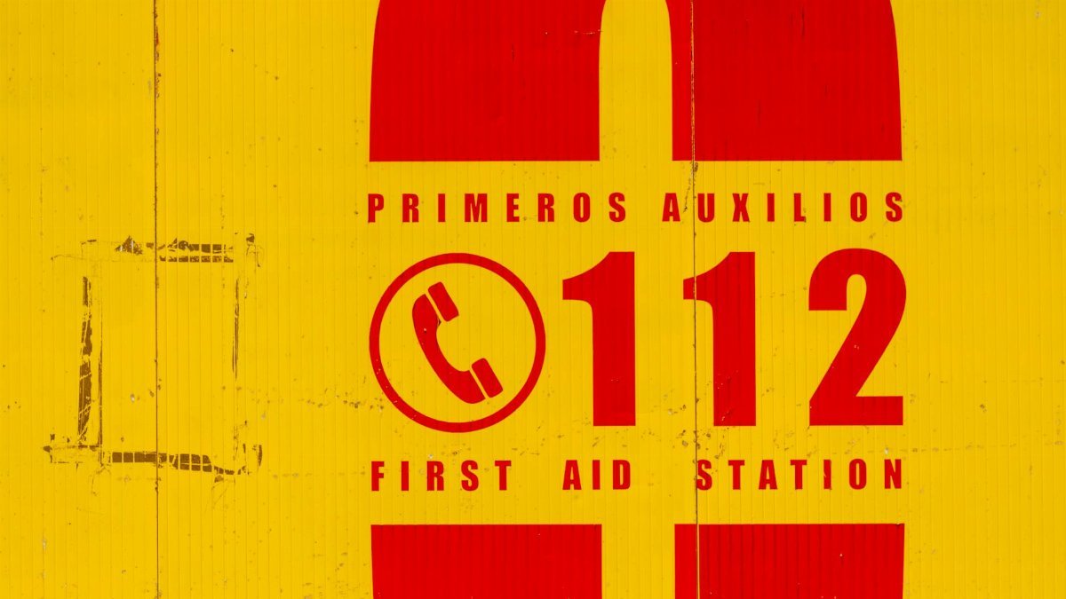 Bright yellow and red emergency sign indicating first aid station. Call 112 for emergencies.