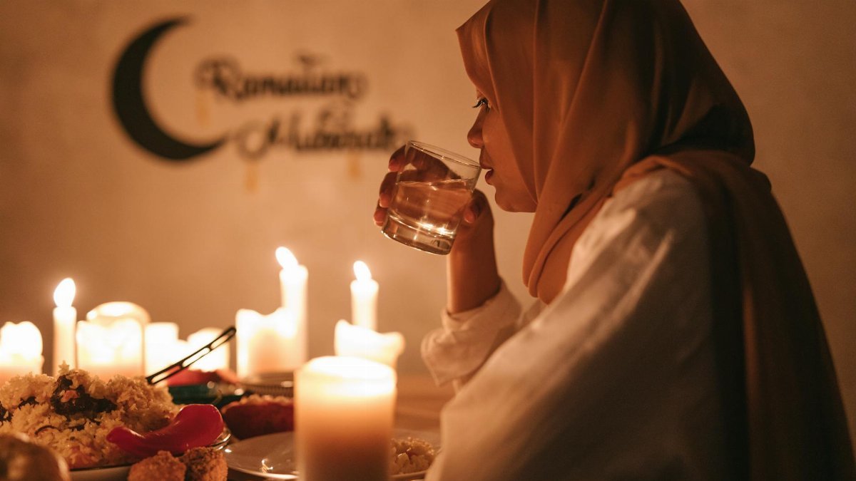 A woman in traditional attire enjoys iftar at a candlelit table during Ramadan, drinking water with a serene backdrop.