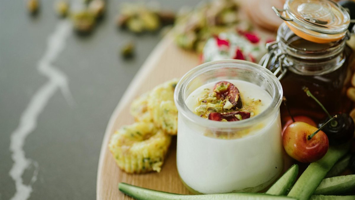 High angle of delicious yogurt with decorative ingredients in glass near cucumber slices and cherries near jar with honey on table in light room
