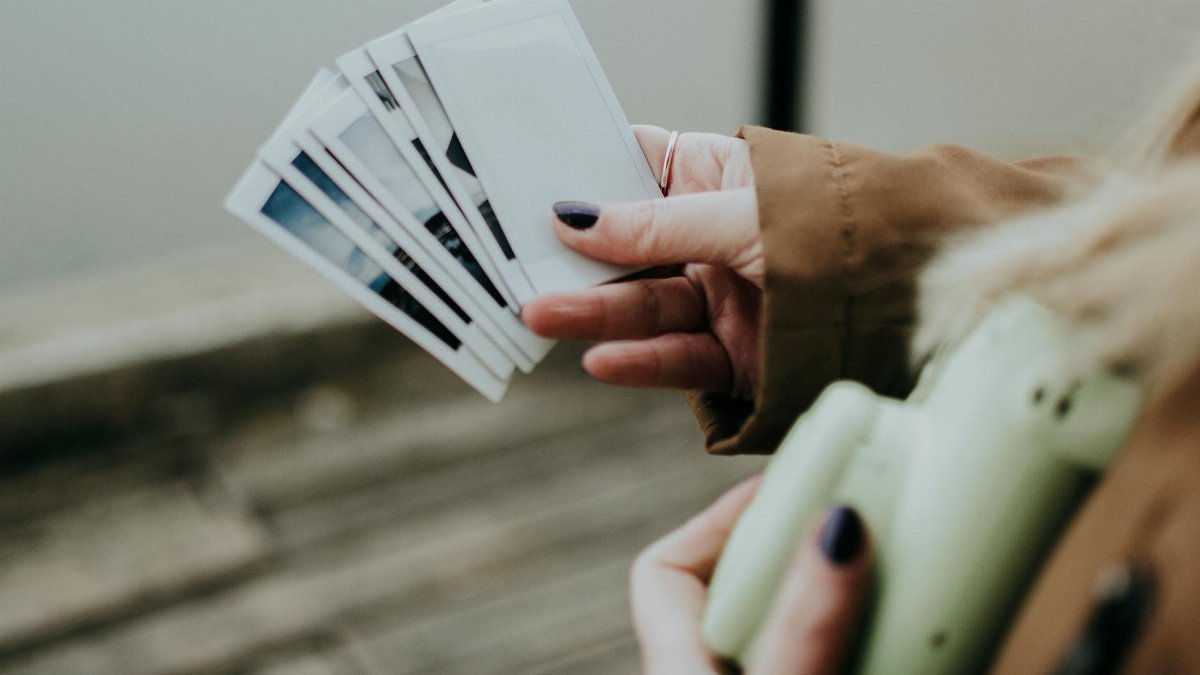 A woman in a brown coat holds a set of instant photos and a camera outdoors.