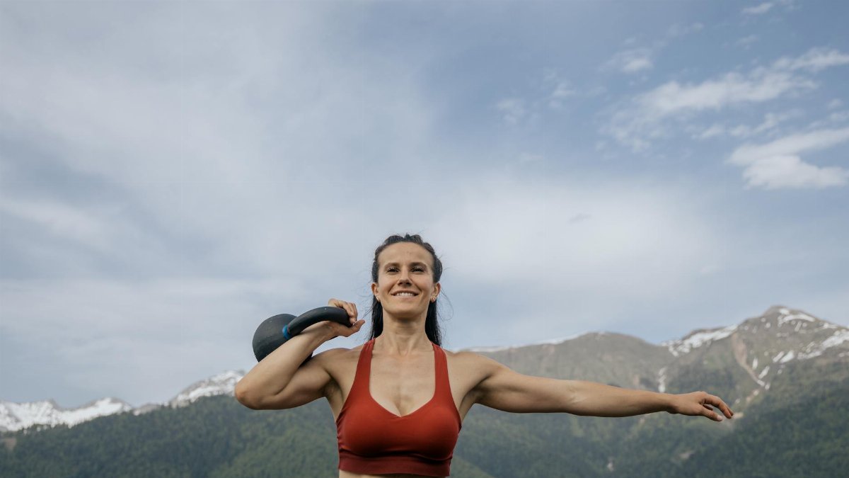 A fit woman in sportswear exercises with a kettlebell in a scenic mountain landscape.