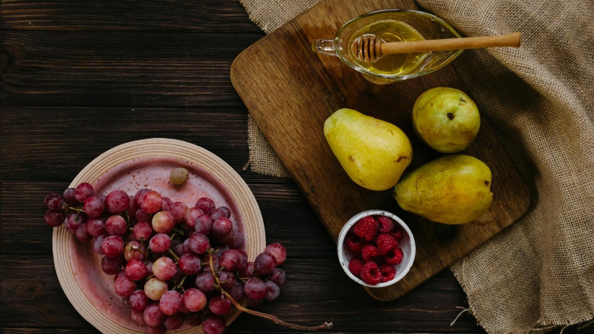 A flat lay of grapes, pears, raspberries, and honey on a wooden board, enhancing healthy eating vibes.