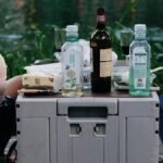 Two people enjoying a lakeside picnic with drinks and snacks on a summer day.