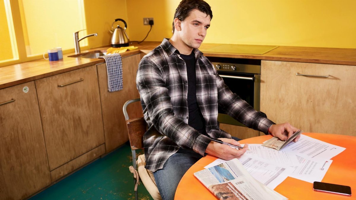 Man sitting in kitchen reviewing bills and a check with smartphone nearby.