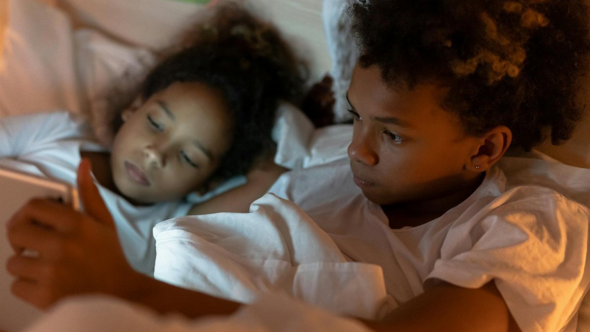 Two children in pajamas lying in bed, engaging with a tablet screen under cozy lighting.