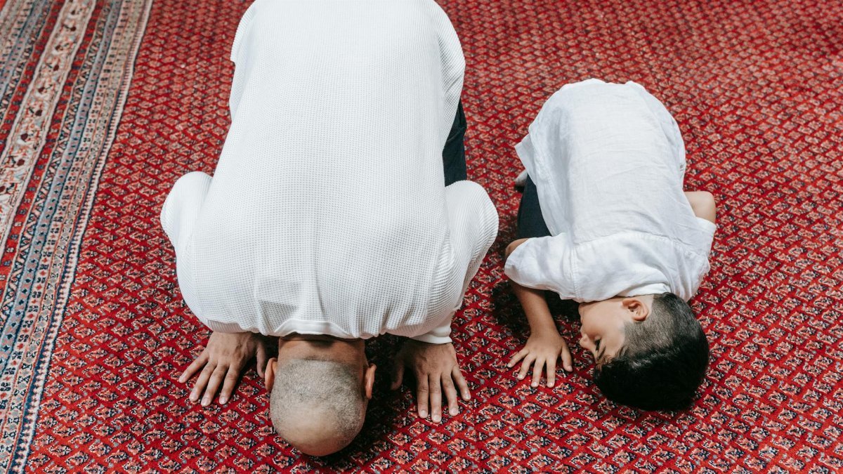 A father and son kneeling in prayer on a beautifully patterned red carpet, symbolizing religious devotion.