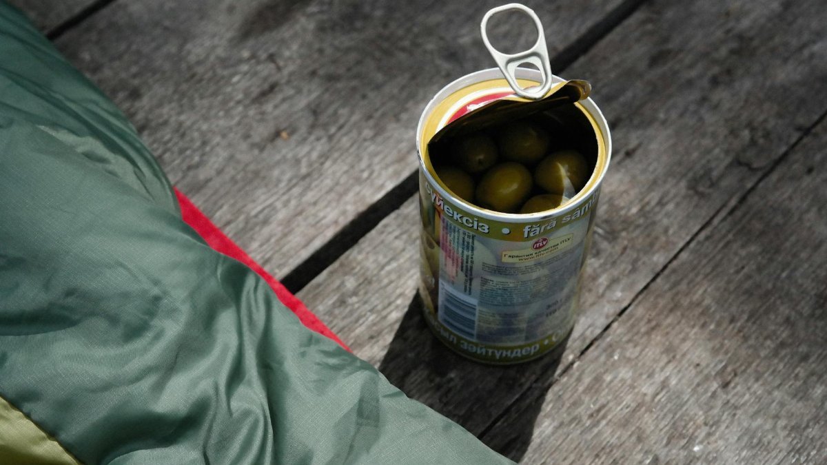 A can of olives opened on a rustic wooden surface with sunlight casting shadows, suggesting an outdoor setting.