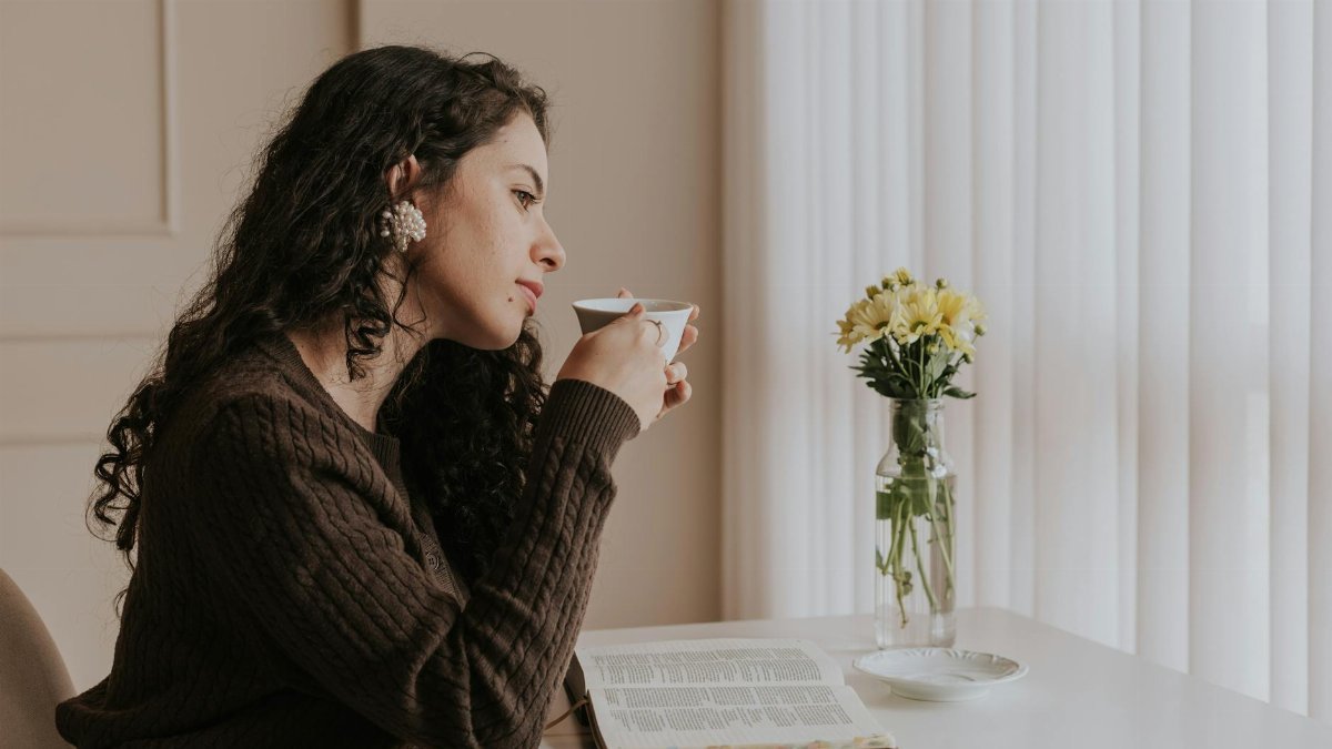 A woman relaxes with tea and a book by a bright window, embracing a peaceful moment.