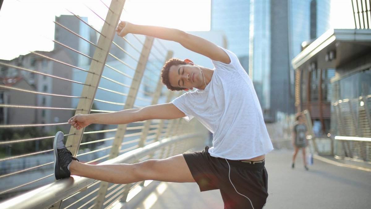 Calm focused African American male athlete with eyes closed in earphones and sportswear leaning on metal fence and doing side bend exercise while listening to music and stretching body on street against blurred urban environment in sunny day