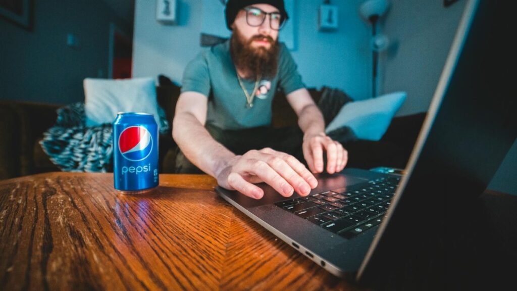 Bearded man wearing beanie, typing on laptop with soda can in cozy workspace.