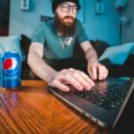 Bearded man wearing beanie, typing on laptop with soda can in cozy workspace.