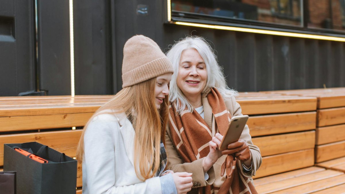 A senior woman sharing a moment with a teenager while looking at a smartphone outdoors on a wooden bench.