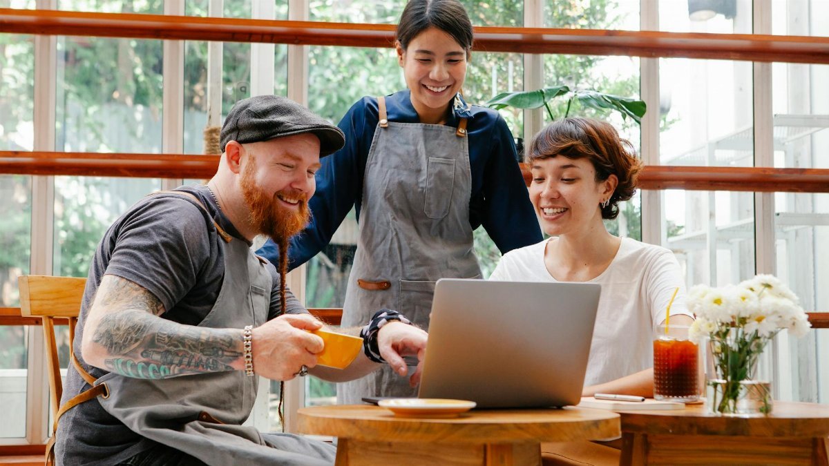 Positive coworkers in aprons and casual outfit using computer in cafeteria at table with coffee cup and glass of drink in day