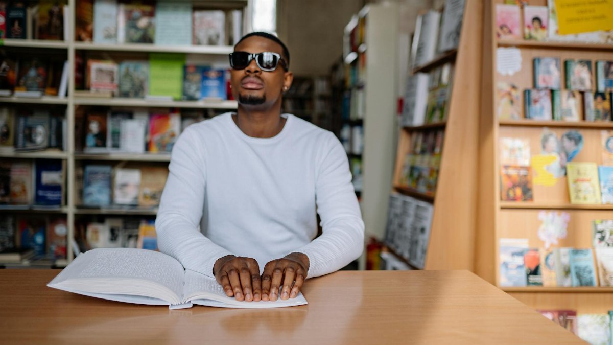 Blind man in white sweater reading Braille at a library desk, enhancing education and accessibility.