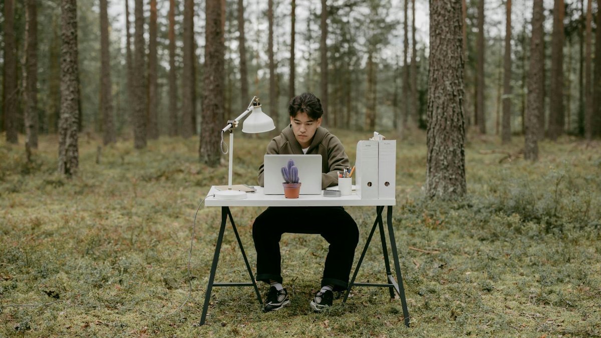 A young man works on a laptop at a desk in a serene forest, embodying the digital nomad lifestyle.