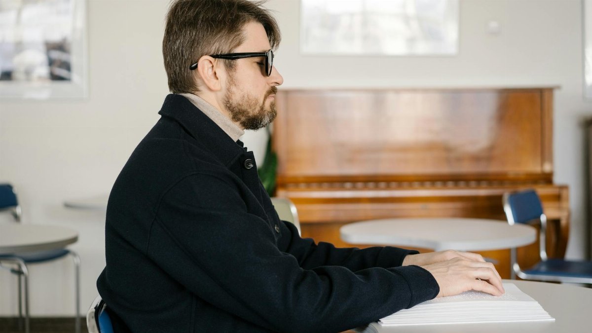 A visually impaired man reading Braille at a table in a warmly lit room with a piano in the background.