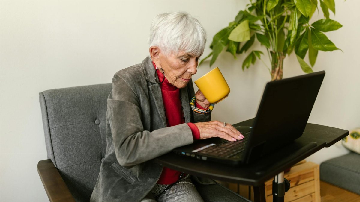 Elderly woman in gray blazer using laptop while drinking coffee, sitting at a home office desk.