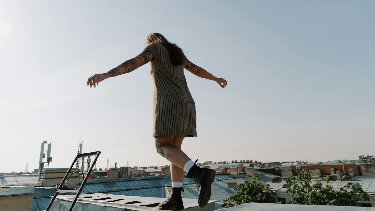 Young woman in summer dress balancing freely on a rooftop edge, exploring urban cityscapes.