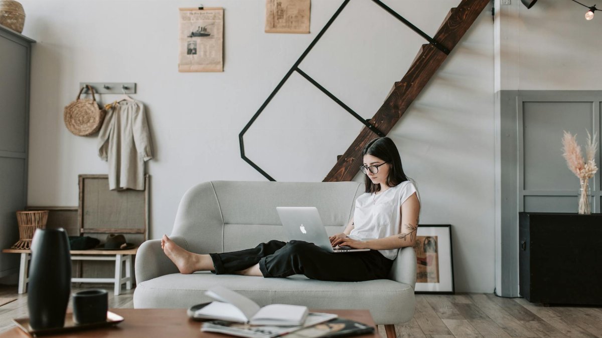 Positive female with tattooed arms wearing casual clothes and eyeglasses reading netbook while sitting barefoot on comfortable sofa behind table with books and magazines in modern apartment with minimalist interior in eco style during free time