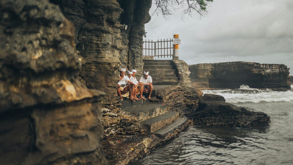 Group of men in traditional attire sitting by a coastal cliff with waves crashing in the background.