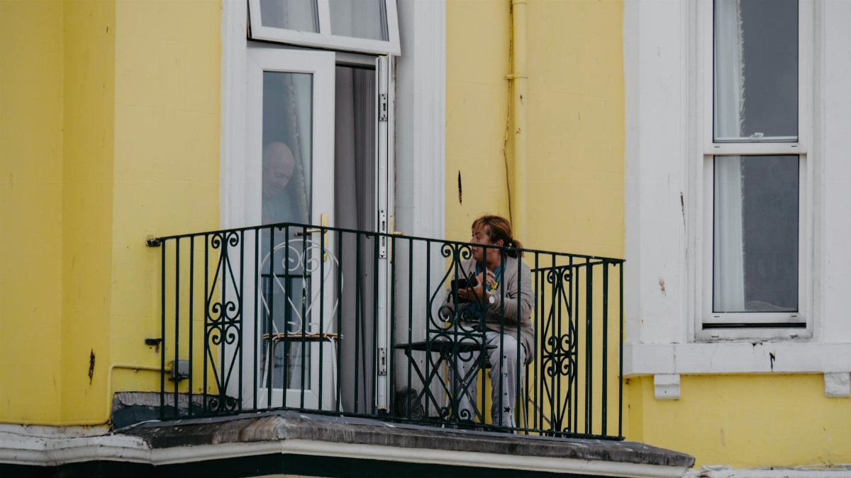 A woman sitting on a balcony of a yellow building, using her smartphone in an urban setting.