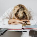 Tired woman in white shirt resting her head on a desk filled with work materials.