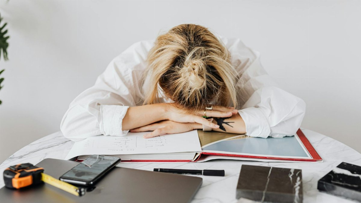 Tired woman in white shirt resting her head on a desk filled with work materials.