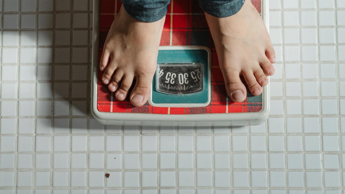 From above crop anonymous barefoot child in jeans standing on weigh scales on tiled floor of bathroom